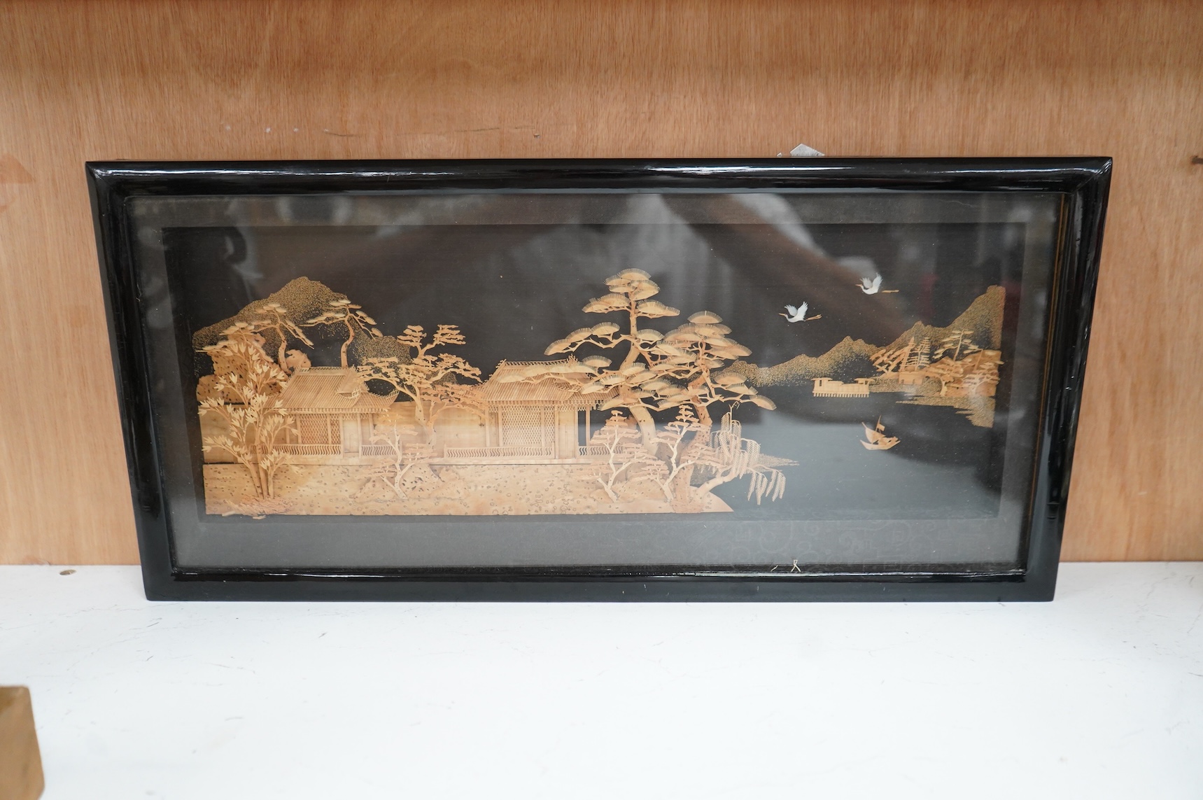 A Chinese framed, intricate relief cork landscape carving, of a Pagoda amongst trees by a river with junks and birds above., 41cm x 15cm